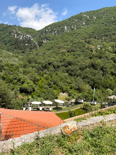 The scene depicts a landscape in Krapina, Montenegro. A steep, tree-covered mountain occupies the left side of the frame, extending upwards into a blue sky with white clouds. On the right side, a red-tiled roof dominates the foreground, with a stone wall covered in vegetation extending upwards from it. Beyond the wall, a green pond is visible, bordered by a black metal fence. Several white parasols are positioned next to the pond, suggesting a recreational or dining area. No people are visible in the image.