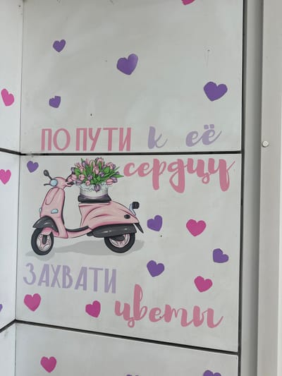 The image shows a white metallic surface, possibly a locker or a cabinet, with painted pink and purple decorations. The decorations include several hearts of varying sizes and two main phrases written in stylized Cyrillic script. The top phrase reads "ПО ПУТИ К ЕЁ" in pink and purple. Below this, a pink cartoon scooter with a basket of tulips is depicted. The bottom phrase reads "ЗАХВАТИ ЦВЕТЫ" also in pink and purple. The overall aesthetic is whimsical and cheerful, with a clear theme related to flowers and romance. The image was taken in Lytkarino, Russia.