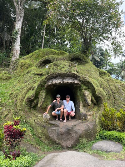 Two men are positioned inside the opening of a large, moss-covered stone structure resembling a monstrous face, likely a cultural or tourist site. The first man, closest to the foreground, wears a dark green shirt, blue shorts, sunglasses, and a hat. He has his arm extended, touching the rock formation. The second man, behind the first, wears a light blue shirt, red shorts, and sunglasses. They are both squatting inside the dark opening. The surrounding area is lush with dense tropical vegetation, including trees, plants, and grass. The image is taken at Goa Gajah Temple in Tegal Suci, Indonesia.