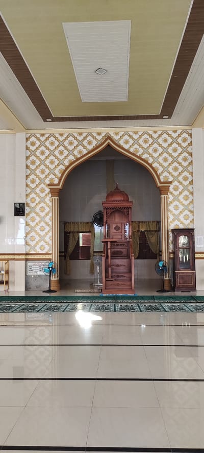 The image shows the interior of a mosque in Peurupok, Indonesia. The focal point is a wooden minbar, a raised pulpit, situated within an arched alcove decorated with a gold and white patterned wallpaper. The minbar is intricately carved and has multiple steps leading up to a platform. In front of the minbar, a green prayer rug with a mosque motif is visible. To the left of the minbar, a black oscillating fan stands on the floor, and to its right, a white oscillating fan is placed on a step. A wooden cabinet with glass doors, possibly for storing religious texts or artifacts, is positioned to the right of the alcove. The floor is made of large, light-colored tiles with dark grout lines. Above, the ceiling is a combination of light yellow and white panels, with brown decorative trim. Natural light streams in from unseen windows, creating bright reflections on the polished floor. The overall scene depicts a serene and well-maintained prayer space within the mosque.
