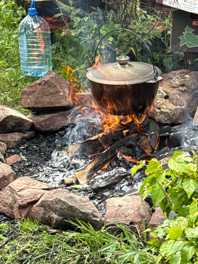 A campfire is burning in a stone fire pit, with a pot suspended over the flames. Next to the fire pit, on the grass, is a large, clear plastic bottle filled with a blue liquid. The surrounding environment appears to be natural, with green foliage and rocks. The image is taken from a medium shot, capturing the campfire and its immediate surroundings. The location is the city of Nizhnyaya Teberda, Russia.