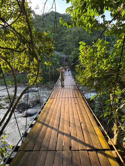 A person is lying down on a wooden suspension bridge, basking in the sun. The bridge spans over a rocky river amidst a lush green forest. Sunlight filters through the dense foliage, casting shadows across the wooden planks. The scene suggests a serene and relaxed atmosphere, likely during daytime with clear weather. The person's posture and the peaceful setting evoke a sense of tranquility and enjoyment of nature. The location, Ranong, Thailand, hints at a tropical environment.