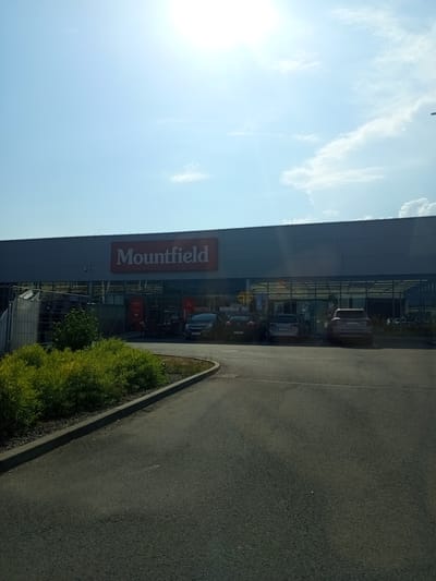 This is a rotated image of the exterior of a Mountfield store in Zvolen, Slovakia. The store features a large glass facade with the red Mountfield logo prominently displayed. Several cars are parked in front of the store, visible through the glass. To the right of the store is a paved area, likely a parking lot or walkway, and a strip of green grass and bushes. The sky is a clear blue with some scattered clouds, and the sun is shining brightly, suggesting it is daytime. There is no visible activity or people in the image. The overall scene is a typical retail environment on a sunny day.