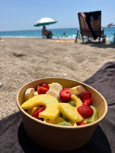 The image shows a fruit salad in a bowl, containing strawberries, banana slices, pineapple chunks, and kiwi pieces, resting on a sandy surface near a dark-colored towel. In the background, out of focus, are beach chairs, people, a beach umbrella, the sea, and a clear sky, suggesting a beach setting. The photo was taken in the city of Municipal Unit of Lefkada, Greece.