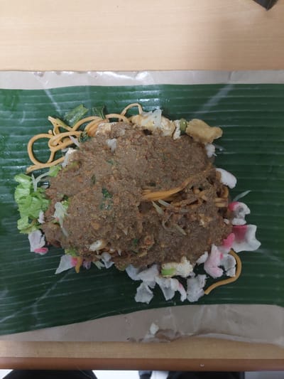 A top-down, close-up shot captures a plate of Indonesian food, likely Pecel, presented on a banana leaf, which is resting on a thin layer of brown paper. The food is a medley of vegetables, noodles, and pieces of tofu or tempeh, all covered in a thick, brown peanut sauce. Accompanying the main dish are crispy crackers, some white and some with pink edges. The food is arranged centrally on the banana leaf, which itself is placed on a light brown wooden surface, hinting at a casual dining setting. The lighting suggests an indoor environment, possibly a restaurant or a food stall in the city of Pematang Siantar, Indonesia.
