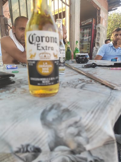 In Dorkovo, Bulgaria, at a roadside establishment, a group of men are gathered around a table. In the foreground, a Corona Extra beer bottle is prominently displayed, its label partially visible. To the left, a man with a white towel draped around his neck is seated. Further back, other individuals are visible, including one wearing a light blue polo shirt. Behind them, a refrigerator stocked with beverages stands against a brick wall, suggesting a place of business. The overall setting appears to be outdoors, under a covered area, with an informal gathering taking place.