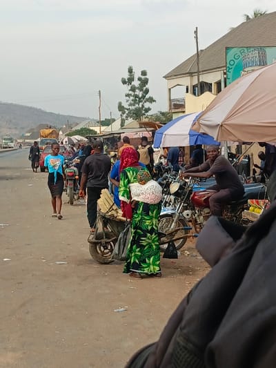 A busy street scene shows a group of people and motorcycles clustered near the roadside. A woman in a green and black patterned dress carries a child on her back and has produce attached to a small wheeled cart. Another woman in a blue top and black skirt walks along the road. A man sits astride a motorcycle. Structures and a sign reading "PROGRESS" are visible in the background, with hills in the distance. The light is muted, indicating overcast conditions.