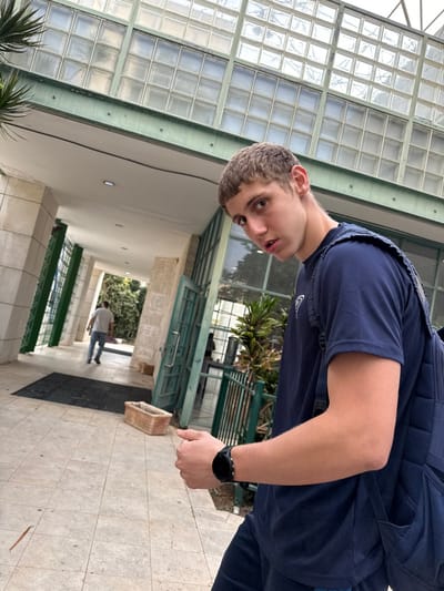 The image depicts an outdoor scene at a building entrance in Ness Ziona, Israel. In the foreground, a young male subject with short brown hair and light skin looks directly towards the camera with a slightly open mouth. He wears a dark blue short-sleeved t-shirt with a light-colored triangular logo on the left chest, dark blue shorts, and a dark blue backpack with a grid pattern. A black smartwatch is visible on his left wrist, and his left hand is partially clenched.

The background features a modern building with a multi-pane glass block facade on its upper level, above a green metal frame. Below this, a covered walkway extends into the distance, supported by light-colored pillars. The walkway floor is paved with light-colored square tiles. To the left, a portion of a palm-like plant is visible. To the right of the walkway entrance, behind a green metal railing, there is a potted plant with large green leaves. Further down the walkway, a second male subject, wearing a light-colored t-shirt and blue jeans, walks away from the camera. A black doormat lies on the walkway, and a rectangular light brown planter box sits on the tiled ground. Green-framed glass doors are visible, leading into the building. The scene is illuminated by natural daylight.