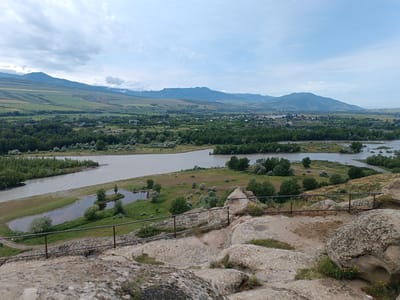 This image captures a panoramic view of the ancient cave city of Uplistsikhe, Georgia, from a rocky, elevated vantage point. In the foreground, rough, weathered rocks form the immediate landscape, with a rusted metal railing in place for safety, suggesting a viewing platform. Beyond the rocks, a lush green valley unfolds, dominated by a wide, meandering river with a light brown, somewhat murky hue.
The riverbanks are lined with dense, verdant trees, creating a sharp contrast with the arid appearance of the foreground rocks. In the middle ground, the river curves, creating small islands and inlets. Scattered across the valley floor are scattered buildings, indicating a village or small town nestled amongst the trees and fields.
Further in the distance, a range of majestic mountains forms the backdrop. The sky above is a mix of soft blue with scattered, wispy white clouds, suggesting a partly cloudy day. The overall impression is one of historical significance and natural beauty, showcasing the integration of human habitation with the surrounding landscape. There are no visible people or animals in the frame.