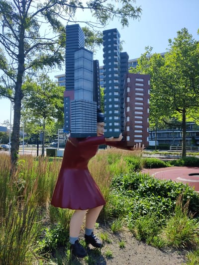 This is a medium shot, eye-level, outdoor photograph taken on a sunny day in the city of Rijswijk, Netherlands, at the 'Sculpture Garden of Rijswijk' located near the city hall. In the foreground, a life-sized, colorful sculpture of a woman is depicted from the waist down, wearing a maroon dress and black sneakers. She is holding a collection of miniature skyscrapers above her head with both hands. The sculpture is situated amidst tall, dry grasses and green foliage. In the background, the scene transitions to a street with cars driving by and modern, multi-story buildings, some of which appear to be offices or apartments. Trees with green leaves are scattered throughout the background, some are quite large and casting shadows. A red circular track, possibly for sports or recreation, is visible on the far right. The sky is clear and blue.