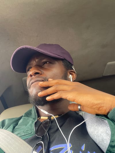 A man is in a car in Sobon-Gida, Nigeria. He is wearing a purple baseball cap, a dark t-shirt with a pattern, and a green jacket. He has earbuds in and sunglasses next to him. He is wearing a seatbelt and a ring on his finger. His hand is partially covering his face. The car interior is visible.