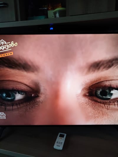 The image shows a close-up of a television screen displaying a close-up of a woman's eyes. The screen also shows text that appears to be in Russian, with "КИЗОВО" and "ПРЕМИУМ" visible. In the lower left corner of the screen, a logo that appears to be "НТВ HD" is visible. Below the television, a white remote control is on a wooden surface. The background of the image shows a shelf with various items, including what appear to be food containers. The lighting suggests the room is dimly lit, with the television screen being the primary light source. There is no indication of the city of Jelgava, Latvia in this image.