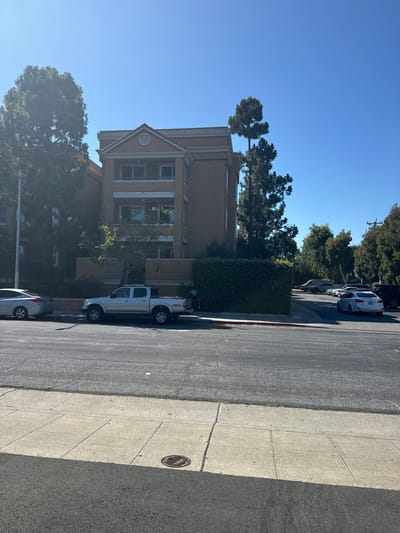 In the city of San Mateo, California, United States of America, a sunlit street scene is captured. The image features a light brown, multi-story building with a pitched roof, flanked by lush green trees and well-maintained hedges. In front of the building, a silver Toyota Tacoma pickup truck is parked. A silver sedan is parked to its left. The foreground shows a paved road with a sidewalk. The sky is clear and blue, indicating a bright day.