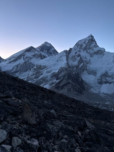 The image presents a majestic, high-altitude mountain landscape in Khumjung, Nepal, likely depicting a view of the Himalayas. The scene is dominated by towering, snow-capped peaks under a clear, pale blue sky.
The upper and left sections of the frame show a serene, gradient light blue sky, free of clouds, indicating clear weather conditions. The lighting is soft, suggesting either early morning or late afternoon, as evidenced by the cool tones and the way light and shadow define the mountain contours.
In the mid-ground and extending into the background on the left, an immense range of snow-covered mountains rises dramatically. These peaks are rugged and sharp, characterized by extensive fields of white snow and ice interspersed with dark, exposed rock faces. The intricate textures of the mountains reveal deep crevices, rocky outcrops, and varying snow patterns, emphasizing their geological grandeur and immense scale.
The foreground, situated in the lower-right portion of the image, consists of dark, rough, and angular rocky terrain. These rocks appear to be close to the viewer, forming a shadowed slope or a collection of boulders. Their dark, irregular shapes and coarse textures provide a strong contrast to the bright snow and sky, anchoring the composition and suggesting a steep, raw environment.
No people, animals, or artificial structures are visible, highlighting the remote and untouched nature of this wilderness. There is no visible text within the image. The overall impression conveyed is one of profound natural beauty, immense scale, and serene isolation within a cold, high-altitude setting.