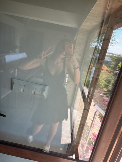 In a reflection on a glass surface, a young woman stands indoors, holding an orange smartphone and making a peace sign with her left hand. She is wearing a black dress and white shoes. In the background, a light switch panel with five toggles is mounted on a light-colored wall. The reflection also shows a glimpse of a living room with white furniture. The glass surface provides a view of an outdoor scene with a lush garden, flowering plants, and a blue sky. A metal railing and a brick wall are also visible in the outdoor reflection. The scene appears to be within a residential building in Aytos, Bulgaria, given the context of the provided information.