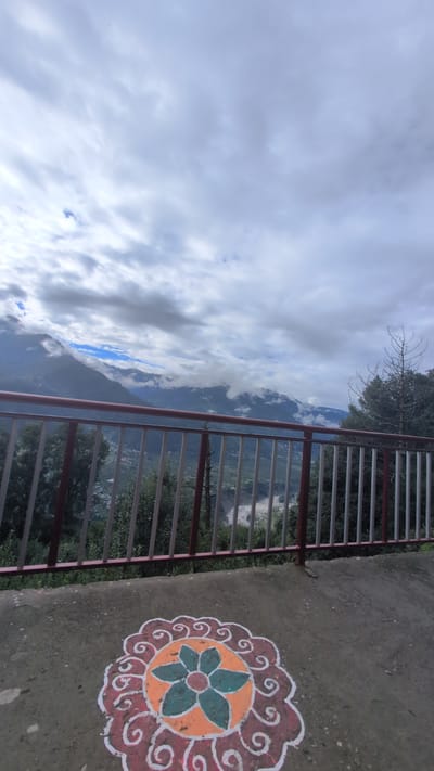 The image depicts a scenic view of a mountainous landscape, likely in or near Pangan, India, under a cloudy sky. In the foreground, a concrete surface features a colorful rangoli, a traditional Indian folk art. The rangoli is circular with intricate patterns in orange, white, and green. Beyond the rangoli, a metal railing with red and white bars separates the foreground from a valley below. This valley contains scattered buildings, indicating a settlement, and a river or water body that appears murky or muddy. Lush green trees and foliage are visible on the slopes of the mountains, which are partially obscured by clouds and mist. The overall impression is one of natural beauty and a glimpse into rural Indian life. There are no people or animals visible in the frame.