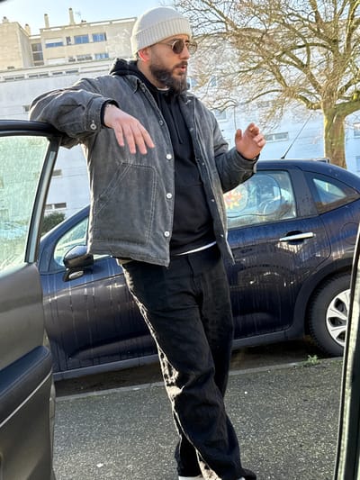 A male subject is depicted standing outdoors on a sunny day in Hérouville-Saint-Clair, France. He is positioned in the midground, leaning with his left arm on the open driver's side door frame of a dark blue or black hatchback car parked on an asphalt surface. The man wears a light beige knit beanie, round-frame sunglasses, a dark grey distressed-look jacket over a black hooded sweatshirt with a sliver of a white undershirt visible at the waist, and black trousers; a silver bracelet is visible on his left wrist. His right hand is raised slightly, fingers curled, as if gesturing or holding an object, and a faint wisp of vapor rises near his face. In the background, a bare deciduous tree stands behind the car, next to several multi-story concrete apartment buildings under a clear sky. The interior door panel of another vehicle is partially visible in the extreme foreground, to the left and right.