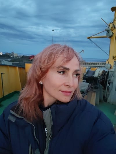 A medium shot captures a woman with pinkish-blonde hair and a dark blue jacket looking to her right. She wears dramatic black eyeliner and a subtle pink lipstick. A heart-shaped earring adorns her left ear. She stands on a deck, likely on a ship or industrial platform, with yellow railings and machinery visible in the background. The sky above is a dusky blue with scattered clouds. In the distance, a coastal industrial area with a red truck and piles of what appear to be coal or gravel is visible. The setting suggests an outdoor, possibly maritime or industrial, environment in Shcheleiki, Russia, under twilight conditions.