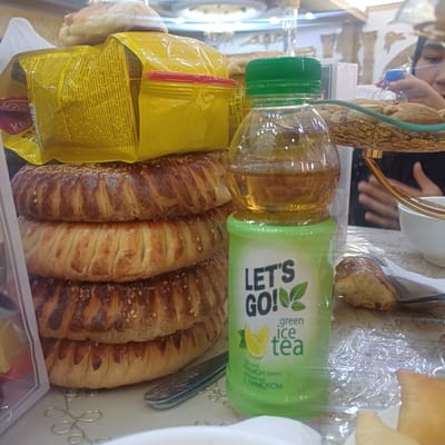 The image captures an indoor scene with a table setting, likely in Osh City, Kyrgyzstan. In the foreground, a stack of four round, baked goods, possibly samsa or flatbread, topped with sesame seeds, are prominently displayed. To the right of the baked goods, a clear plastic bottle of "LET'S GO! green ice tea" is visible, its contents a pale amber liquid. Behind the bottle, a golden decorative tray holds a pile of unshelled peanuts. Further in the background, a person's hand is visible reaching for a blue-capped bottle, and another person's face is partially obscured. The table is covered with a patterned tablecloth, and various other objects, including a white bowl and what appears to be more baked goods, are present. The overall impression is of a casual gathering or meal.