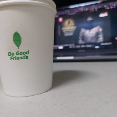 A white disposable paper cup, adorned with a green leaf logo and the phrase "Be Good Friends," rests on a light-colored, speckled surface in 김포시, South Korea. In the blurred background, a laptop screen displays a figure wearing a dark hood and a camouflage-patterned jacket, alongside a golden emblem and obscured text. The scene implies an indoor setting, likely a desk or table, with the cup positioned in the foreground and the laptop screen providing ambient light. No individuals are visible.