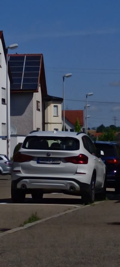 The image shows a white BMW X3 parked on a paved road in Schwaikheim, Germany, during the daytime. The car is facing away from the camera, displaying its rear bumper, taillights, and license plate which appears to read "BB-RI-1527". To the right of the white BMW, a dark-colored SUV, possibly a BMW as well, is partially visible. In the background, a building with a dark-tiled roof and solar panels is situated to the left, with streetlights visible along the road. The sky is clear blue with some faint clouds. The environment suggests a residential or commercial area.
