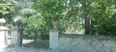 The image depicts an outdoor scene, likely in San Rafael, Spain, featuring a stone wall and gate. The wall is constructed from irregularly shaped gray stones, forming a rustic barrier. To the left of the wall, there is a gate composed of vertical black metal bars. Two stone pillars, topped with spherical ornaments, flank the gate. Behind the wall and gate, dense green foliage, including trees and bushes, creates a natural backdrop. Sunlight filters through the leaves, casting dappled shadows. A paved surface, possibly a sidewalk or road, is visible in the foreground. The scene conveys a sense of established property, likely an entrance to a garden or estate.