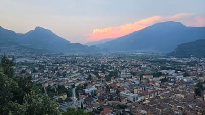 This is a high-angle, wide shot of Riva del Garda, Italy, taken during the twilight hours. The sky is a gradient of soft blues and pinks, with a dramatic band of illuminated clouds stretching across the horizon, catching the last rays of the setting sun. In the foreground, to the left, is a dense cluster of dark green trees and foliage.

Below the trees, the city of Riva del Garda unfolds, a tapestry of terracotta rooftops and buildings in muted earth tones, interspersed with green spaces. The architecture appears to be a mix of older, classical styles and more modern structures. A paved road curves through the city, with cars visible on it and in parking areas. Streetlights are beginning to illuminate the road and some buildings, indicating the transition from day to night.

In the background, majestic mountains rise, their peaks softened by the dusk and atmospheric haze. The slopes of the mountains are covered in vegetation, varying in shade from dark green to a hazy blue. The overall impression is one of a peaceful, scenic, and historic Italian town nestled within a dramatic natural landscape. There are no people visible in the image.