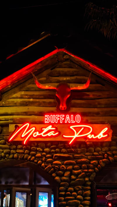 The image depicts the exterior of "Buffalo Mota Pub" in Du Già Commune, Vietnam, at night. The pub features a log cabin facade topped with a roof outlined in red neon. A red buffalo skull is mounted centrally below the roof's peak. A neon sign that reads "BUFFALO Mota Pub" is placed on a sign-board attached to the logs. The lower portion of the building is constructed of stone. Light shines through the windows of the entrance.