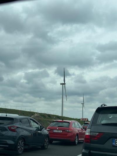 The image shows a traffic jam on a road near a wind farm. The sky is overcast and gray. Several cars are visible, including a black SUV, a red sedan with license plate LL 262 AD, and a dark gray hatchback. A road sign with the number 85 is also visible. The location is near wind turbines, but not explicitly identified as Kisigmánd, Hungary.