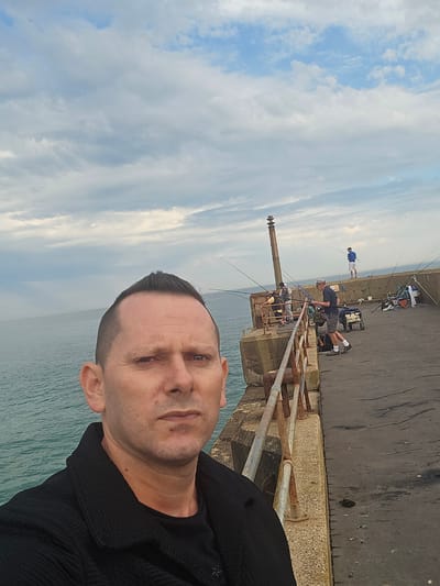 A man is taking a selfie on a pier, with the sea and a cloudy sky in the background. He is wearing a black jacket over a black t-shirt. The pier is made of concrete and has a rusty metal railing. Several other people are visible on the pier, fishing. One person is standing on the far end of the pier, and others are scattered along the length, with fishing rods set up. There are also some fishing equipment and carts visible. The location is on the pier in Shoreham-by-Sea, in the borough of Adur, United Kingdom.
