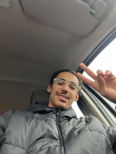 The image depicts a male individual inside a vehicle, reportedly located in Eckbolsheim, France. The subject, positioned centrally and viewed from a low angle, has light-brown skin, dark hair styled in dreadlocks or braids, and a visible mustache. He wears clear-framed eyeglasses and exhibits dental braces on his upper teeth, with a slight upward turn at the corners of his mouth. He is dressed in a black, zippered puffer jacket; a partial 'Adidas' logo is visible on the left chest area. To the right of the subject's head, a hand displays an upright 'peace' sign gesture (V-sign). The vehicle's interior components are visible, including a beige-grey headliner, a partially obscured sun visor, and a dark A-pillar section with two circular caps. An overcast sky is visible through the vehicle's window on the right side of the frame.