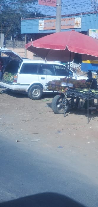 The image captures a vibrant roadside scene during what appears to be a sunny day. The primary focus is a white van or minibus parked on a light brown, unpaved surface, likely a dirt road or shoulder.

Attached to the side of the van, a large, two-toned umbrella is open, providing shade. One half of the umbrella is a bright reddish-pink, and the other half is a darker, brownish hue. Behind the van, a makeshift selling stall or cart is present, laden with what appears to be goods, possibly fruits or vegetables, wrapped in brown packaging or sacks. This cart is attached to the rear of the vehicle and has its own visible wheel. A white sign or plate is affixed to the side of this cart, displaying the text "0536830." Another tire is visible on the ground near the front of the van, suggesting a spare or an additional part of the vendor's setup.

In the background, on the right side of the image, a utility pole stands tall. A red and white rectangular banner or sign is attached to this pole, prominently displaying phone numbers: "0799 745 774" and partially visible, "0720 000 000." Beyond the pole and the vehicle, several buildings with light blue corrugated metal roofs and walls are visible, indicating a street lined with shops or residential structures. The bright lighting suggests it is daytime, and the clear sky implies good weather. Some sparse vegetation can be seen towards the horizon. The overall scene depicts a common sight of a mobile vendor operating a stall by the road.