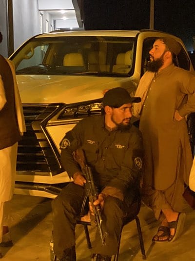 The image shows a man in a dark uniform holding a rifle while seated in front of a white SUV. Another man in traditional clothing stands beside the vehicle. The location is believed to be in قندهار, Afghanistan.