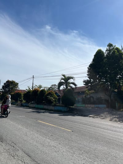 A motorcycle with two occupants, a driver and a passenger, is traveling on an asphalt road in Bukitrata, Indonesia. The passenger, on the back, is wearing a light-colored hooded jacket and a pink and red garment on their lower body. The road has a yellow lane marker and shows signs of wear, with cracks visible. Along the side of the road is a low concrete wall with green and blue trim. Behind the wall are various trees and shrubs, some with reddish foliage. In the background, residential buildings with red tiled roofs are visible, along with utility poles and overhead power lines. A large tree with dense green foliage stands prominently on the right side of the image. The sky is bright blue with scattered white, wispy clouds. The overall scene depicts a sunny day in a suburban or semi-rural area.