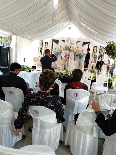 This image depicts an indoor/outdoor wedding event taking place under a large, white-draped tent structure in Juwana, Indonesia.
The central focus is a decorated stage, or *pelaminan*, at the far end of the tent, featuring a bridal couple. The groom wears traditional patterned gold and white attire with a white headpiece, while the bride wears a white dress/kebaya, a white hijab, and a white crown-like headpiece. Both individuals are standing, facing forward, and appear to be holding small, ceremonial objects in their hands. The stage is adorned with white, blue, and light green floral arrangements, and a white backdrop behind the couple displays the words "Taufiq Indah" in stylized script.
To the left of the stage, a male photographer in a dark blue short-sleeved shirt is actively photographing the couple using a DSLR camera. A white table in front of him holds a camera flash and other equipment. To the right of the couple, a male individual in a dark long-sleeved shirt stands, facing them. Further to the far right, a potted plant and another person in dark attire are partially visible. A large, black speaker or sound system unit stands on the far left of the tent.
In the foreground and midground, multiple attendees are seated, facing the stage. They occupy white chairs, most of which are covered with white fabric and tied with white sashes. Several chairs prominently display a circular logo with a red cross symbol centered within a red ring. Visible attendees, mostly viewed from the back, include a man with curly brown hair wearing a dark long-sleeved batik shirt, a man in a black short-sleeved shirt, and a woman in a dark red top. The tent ceiling is extensively draped with white fabric, and multiple modern chandeliers hang overhead. The ground surface appears to be paved or concrete.