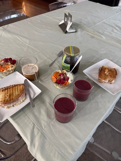 A light green tablecloth covers a dining table in Nogueira e Silva Escura, Portugal, prepared for a meal. The table setting includes two clear glass bowls, each containing a mixture of fruit (raspberries, sliced bananas, diced orange fruit) and granola over a light-colored base. Adjacent to these are two clear glasses filled with a dark red-purple smoothie. Two square white plates each hold a grilled panini-style sandwich with distinct grill marks and visible melted filling; one plate also features a silver knife. A clear glass mug with a handle contains a dark beverage, likely coffee, with a spoon inserted. A glass jar with a labeled lid, displaying yellow and blue accents, holds a dark viscous substance, presumably honey, with a spoon submerged. An additional silver spoon rests on the tablecloth near the jar. A silver napkin holder containing white paper napkins is situated further back on the table. The surrounding environment partially reveals transparent chairs with metal frames and a blurred wooden floor in the background.