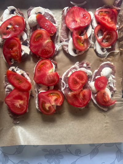 The image shows a top-down view of uncooked food items arranged on a baking sheet lined with parchment paper. The food appears to be meat slices, possibly pork, topped with sliced tomatoes, white onions, and mushrooms. The ingredients are layered on each meat slice, and there are multiple servings arranged in rows. The background consists of the brown parchment paper and a glimpse of a patterned surface at the bottom. There are no people visible in the image. The scene is indoors, likely a kitchen. The location is not specified within the image itself.