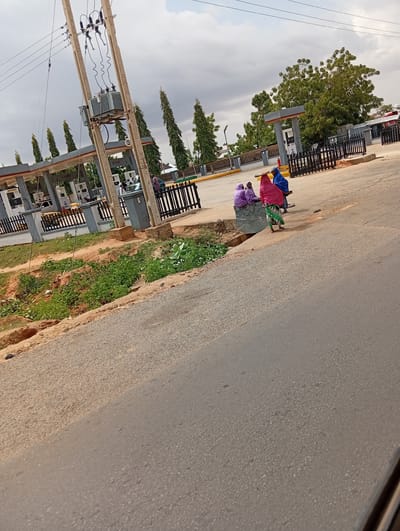 This is a photograph of a gas station in Gadao, Nigeria. In the foreground, a paved road runs diagonally across the frame, with a patch of unpaved, sandy ground and overgrown green vegetation to its left. In the background, the gas station features multiple pumps under a covered canopy. Several people are present, including a group of three individuals wearing colorful hijabs sitting on a concrete structure, and a person in a blue garment standing near them. A tall electrical pole with transformers and wires stands prominently to the left of the gas station canopy. The sky is overcast with grey clouds.