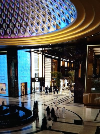 The image captures the interior of a high-end retail space, possibly a luxury shopping mall, with a distinctive architectural design. The ceiling features a large, ornate dome with a mosaic-like pattern of diamond shapes, illuminated by vibrant blue and purple lights, accented by a golden, layered trim. Below the dome, the space opens into a well-lit atrium with a polished floor.
On the left side of the frame, large blue digital screens display branding, with "LOUIS VUITTON" visible on one. Adjacent to these screens are storefronts, including what appears to be a Louis Vuitton boutique with an open entrance. Further into the mall, other shops are visible, with some featuring minimalist designs and illuminated grids on their facades. Trees and decorative plants are interspersed throughout the space, adding a touch of nature.
Several individuals are present in the scene, mostly walking or standing in small groups. Many are dressed in traditional Middle Eastern attire, such as thobes and abayas, suggesting a possible location in a region where this is common. Some are seen near the storefronts, while others are walking across the open floor. In the foreground, a circular water feature or decorative pool is visible, reflecting the ambient light. The overall impression is one of opulence, modern design, and a bustling commercial environment.
This scene is not located in Golbembal, Nigeria, as no information in the image supports this specific location. The architecture, signage, and attire of the people are inconsistent with that region.