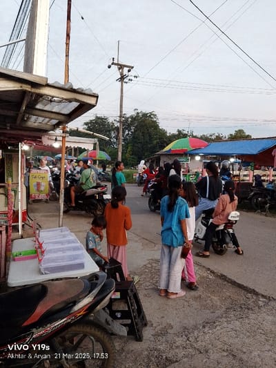 The image shows a street scene in Kecamatan Kroya, Indonesia, with a group of people standing and interacting with each other, possibly near a market stall or a street food vendor. The scene is located on a street with a few parked motorcycles and some small shops on the side. There is a street sign with the text "Teh Coyo" and the price "3,000" visible on the left side of the image. There are several people standing and talking, and some are sitting on motorcycles. The scene is taken on a street, likely a busy thoroughfare. The sky is clear, and the sun is setting. The environment is characterized by a mix of residential and commercial areas, and there is a sense of community in the scene. The image is likely taken in the late afternoon, with the shadows being long and the sky taking on a soft golden hue. The atmosphere is relaxed and casual. The scene is also characterized by a strong sense of local life, and the people appear to be comfortable with their surroundings. The image is well-composed, with the people and objects positioned in a way that creates a sense of depth and interest. The colors are warm and inviting, and the overall effect is one of peace and tranquility. The scene is typical of everyday life in Kecamatan Kroya, Indonesia, and the image captures a moment in time.