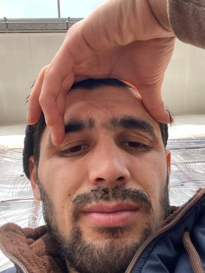 A close-up, slightly angled selfie shows a man with dark hair, eyebrows, and a patchy beard. He has brown eyes and is looking directly at the camera. Part of his hand is covering his forehead. He is wearing a jacket with a brown, possibly sherpa, lined hood and a blue outer layer. The background includes a white wall with a window on the left and a textured, possibly plastic, surface on the right.