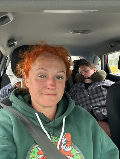 The image depicts the interior of a vehicle, likely a car, where two individuals are present. The person in the foreground, with bright red, curly hair, is looking directly at the camera and appears to be the driver. They are wearing a green hooded sweatshirt with a white drawstring and a dark necklace. Their left shoulder is angled towards the camera. To the right and slightly behind, another individual is visible, sleeping. They are wearing a plaid shirt and have a travel neck pillow around their head, with a strap across their chin. A third person, wearing a hat and a plaid shirt, is partially visible in the background, seated behind the driver. The interior of the car shows the ceiling, a seatbelt, and the back of a headrest. The lighting suggests it is daytime, and the scene appears to be set within or near the city of Tatarstan, Russia.