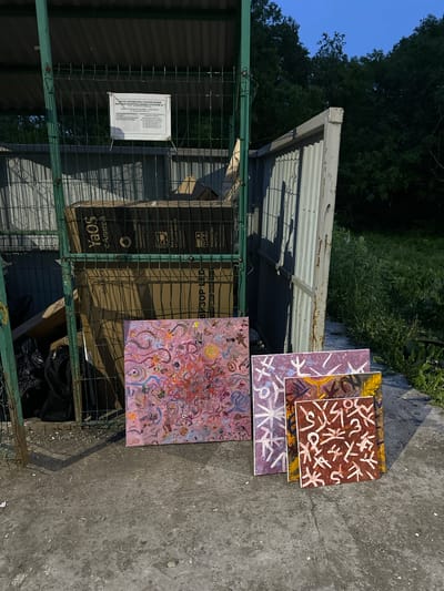 This image depicts an outdoor scene at a garbage collection point in Vilyuchinsk, Russia. The foreground features four paintings leaning against a concrete surface. One painting, in the center, is larger and displays a pink abstract design with swirling lines and various shapes. To its right, two smaller paintings are placed. One is purple with white abstract symbols, and the other is a reddish-brown color with white symbols and yellow accents. A fourth painting, partially obscured behind the others, is also visible.

In the mid-ground, behind the paintings, is a green metal-fenced enclosure with corrugated metal siding. Inside the enclosure, large cardboard boxes are stacked, some appearing to be packaging for electronics. A white sign with Russian text is attached to the fence. To the right of the enclosure, a larger, open corrugated metal structure is present, casting a shadow on the ground. Black trash bags are visible near the base of the green enclosure.

The background consists of dense green trees and foliage, indicating a natural, possibly forested, environment surrounding the urban infrastructure. The lighting suggests it is either early morning or late evening, given the soft, diffused light and clear blue sky. The ground is a mixture of concrete and dirt, with scattered debris. There are no people or animals visible in the frame. The scene appears to be a collection point for large waste, likely in a residential or industrial area of Vilyuchinsk.