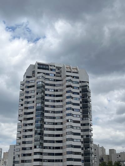 The image shows a modern apartment building in Créteil, France. The building is tall and features a distinct architectural style with many balconies and windows. The facade is primarily light-colored, with darker sections accentuating the balconies and window frames. The building has a stepped or angled design, creating a dynamic silhouette against a cloudy sky. The sky itself is filled with a mix of dark and lighter clouds, suggesting an overcast or possibly stormy day. In the background, other buildings are partially visible, indicating an urban setting. The image is taken from a low angle, looking up at the building. There are no people visible in the frame.