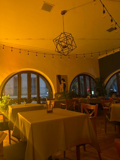 The photograph depicts the interior of a room in ქვედა სამება, Georgia, likely a restaurant or dining area. The room is illuminated with a warm, yellow light from a combination of overhead fixtures: a modern, geometric chandelier, strings of exposed light bulbs, and ambient lighting. The architectural style includes arched windows that offer views of the outside, revealing buildings and greenery. A painting adorns the wall between the windows. The foreground is dominated by tables covered in tablecloths, with wooden chairs arranged around them. The floor appears to be wooden. The composition is oriented vertically.
