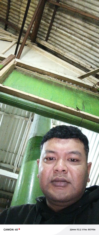 The image shows a close-up portrait of a man with short black hair, captured from the chest up. He is wearing a black hooded sweatshirt and is looking directly at the camera with a neutral expression. The man's background is the interior of a structure with a corrugated metal roof, which is visible through a gap in the wooden ceiling. The image appears to have been captured in the city of Pasuruan, Indonesia, based on the provided coordinates. The location is likely an indoor space with a roof structure visible above. The image was captured with a 22mm lens, f/2.2 aperture, 1/14s shutter speed, and ISO 7106. The photo is taken from a low angle, looking up at the man. The environment appears to be well-lit and the image is in focus. The overall tone of the image is simple and direct, focusing on the man's face and expression. The background details hint at a potentially domestic or work-related environment.