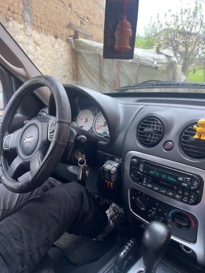 The image depicts the interior of a Jeep, with the driver's perspective of the dashboard, steering wheel, and a portion of the driver's leg. The gear shift and radio are visible on the dashboard. The windshield reveals an outdoor scene of what appears to be a building covered with a plastic tarp and other buildings in Kornitsa, Bulgaria on a rainy day. The windshield is wet, and the wiper blades are visible. A car key is in the ignition. The steering wheel has a Jeep logo. The radio shows a station CD1 T8 is playing, and the time is 21:18.