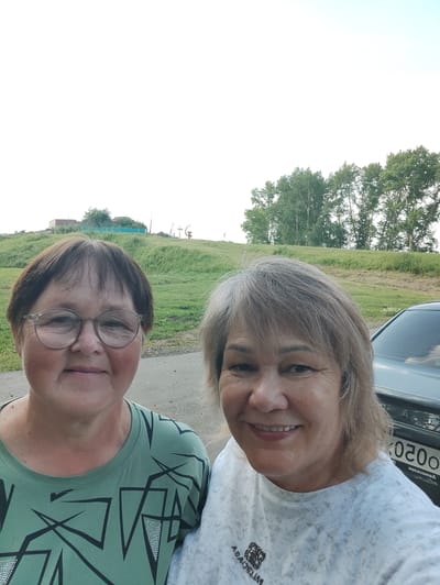 Two women are posing for a selfie outdoors. The woman on the left has short brown hair and is wearing glasses and a green t-shirt with a geometric pattern. The woman on the right has shoulder-length blonde hair and is wearing a light-colored t-shirt with a marbled pattern and a brand logo. A dark-colored car with a license plate partially visible is behind them on the right. The background consists of a grassy hill with some trees and structures at the top, suggesting a semi-rural or park-like setting. The sky is bright and overcast. The location is identified as Karabash, Russia.