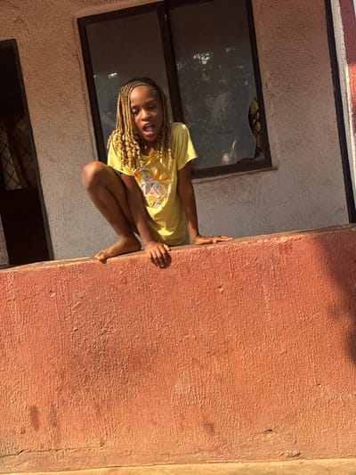 The scene, located in Aguata, Nigeria, depicts a young, dark-skinned girl with long, light brown braided hair. She is positioned in a squat on a prominent reddish-brown textured wall, facing the viewer. The girl wears a yellow short-sleeved t-shirt with a multi-colored graphic design on the front and is barefoot. Her hands are placed flat on the top surface of the wall in front of her. Her mouth is open, and her eyes are wide, directed towards the camera. Behind her is a light-colored, textured building wall featuring a dark-framed window with multiple panes; one pane on the right appears broken or distorted. A partial, dark-framed opening is visible on the far left of the background building. The environment is illuminated by natural daylight, casting some shadows.
