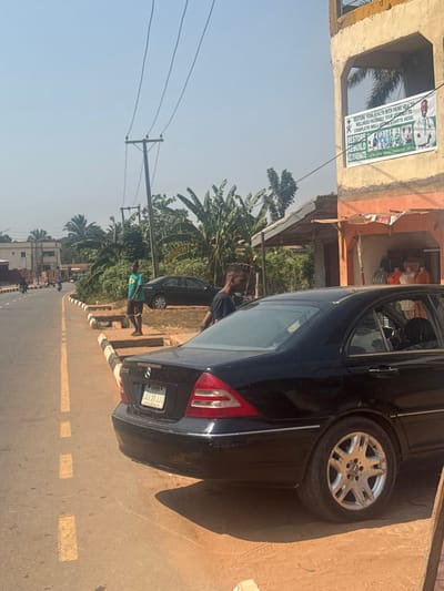 The scene depicts a daylight street view in Aguata, Nigeria, under a clear sky with strong sunlight casting distinct shadows. In the immediate foreground and midground, a black Mercedes-Benz sedan (Lagos license plate "01 JIJ JJJ") is parked off the asphalt road, positioned on a reddish-brown dirt shoulder. A male individual, wearing a dark t-shirt, stands adjacent to the car, partially turned away from the camera. To the left, the road features a solid yellow line on the far edge and dashed yellow markings further inward, bordering a black and white striped curb. A second male individual, wearing a green t-shirt with a yellow emblem and dark shorts, stands barefoot on the raised curb area.

In the midground and background, utility poles with multiple overhead power lines are visible against the sky. Dense tropical vegetation, including large-leafed plants and palm trees, occupies the space behind the parked vehicles. On the right, a two-story concrete building with a weathered facade features an open storefront on the ground floor, revealing several indistinct figures in orange attire inside. The upper floor displays a large banner advertising "Prime Health" wellness packages, with text such as "RESTORE YOUR HEALTH" and "RESTORE REBUILD REJUVENATE," accompanied by images of medical professionals. Further down the road on the left, another dark-colored sedan is parked on a dirt shoulder, partially obscured by foliage, with additional buildings and faint vehicle movements visible in the distance.